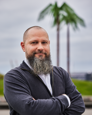 Ein Mann mit Bart steht mit verschränkten Armen vor künstlichen Palmen