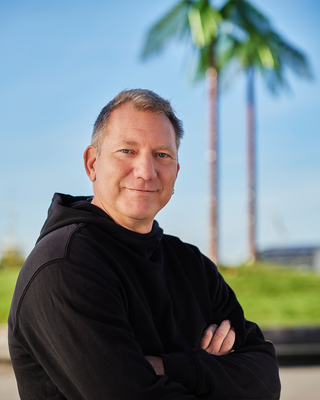 Ein Mann steht mit verschränkten Armen vor künstlichen Palmen und lacht