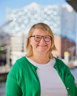 Frau mit blonden Haaren, Brille, weißem Oberteil und grüner Weste steht vor der Elbphilharmonie