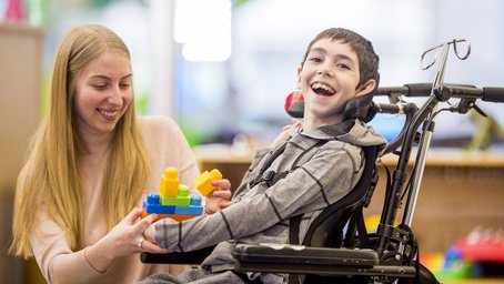 Frau reicht Spielzeug an Jungen im Rollstuhl.